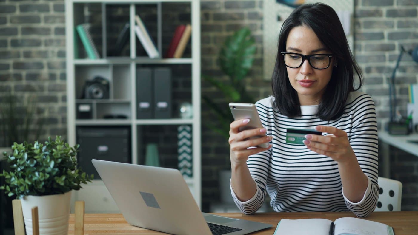 Une femme hésitant, sa carte bancaire à la main, devant son téléphone et son ordinateur
